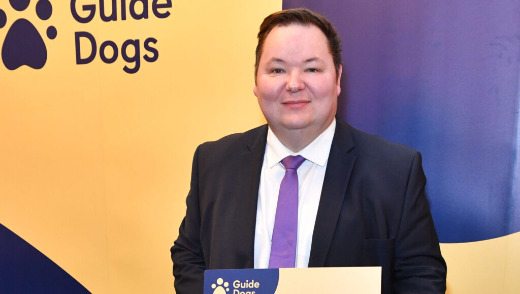 Andrew with a golden retriever trainee Guide Dog holding a sing which reads 